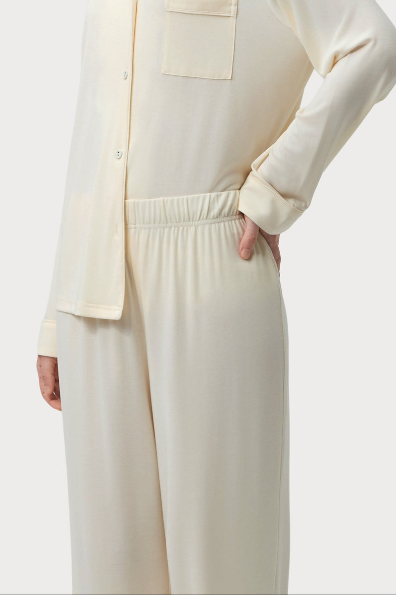 A model wearing a cream-colored two-piece pajama set consisting of a long-sleeve button-up shirt with a chest pocket and matching wide-leg pants, shown against a light gray studio background.